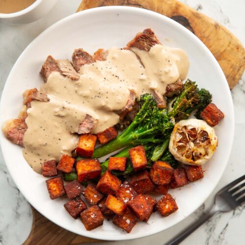 sliced steak with creamy garlic sauce on white plate with potatoes and broccolini